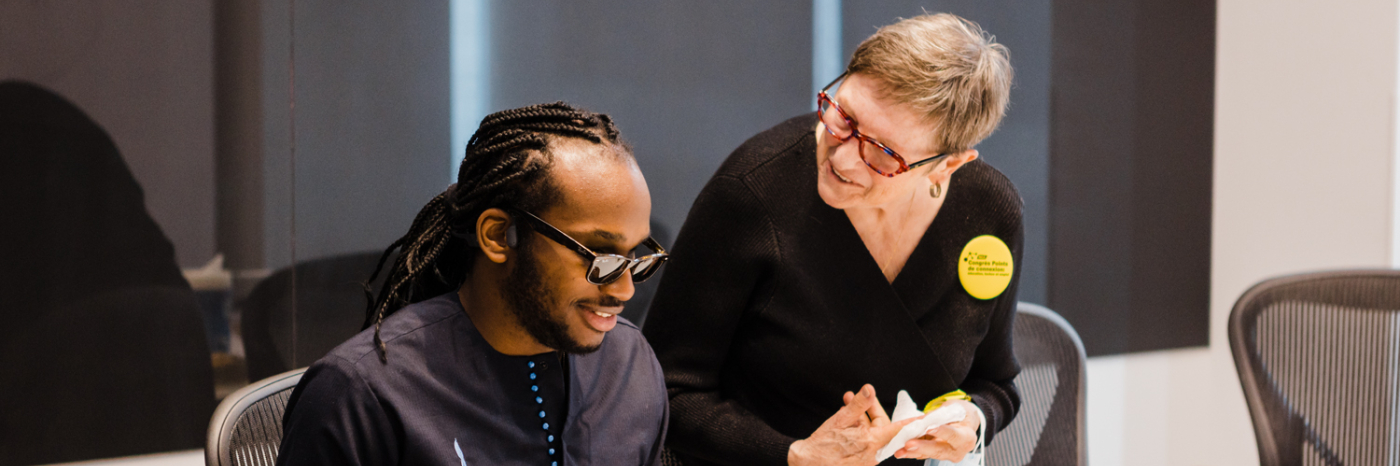 A young black man with sight loss and a middle-aged white woman sit at desk while having a conversation. There are boxed lunches on the table.