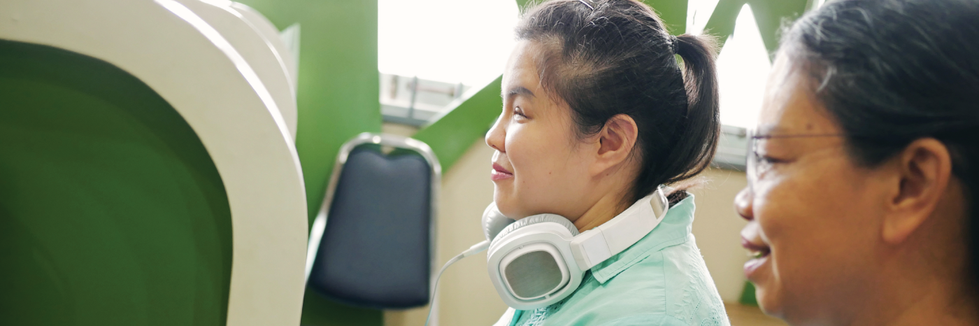 In an office cubicle, a woman sits at a computer workstation and types on a keyboard. A pair of large headphones rest around her neck. Next to her, a colleague sits at another workstation. They are reviewing something work related on the computer monitor. 