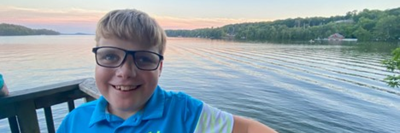A young boy wearing glasses and holding a white cane is leaning against a wood railing. Behind him is a lake with trees on the horizon.