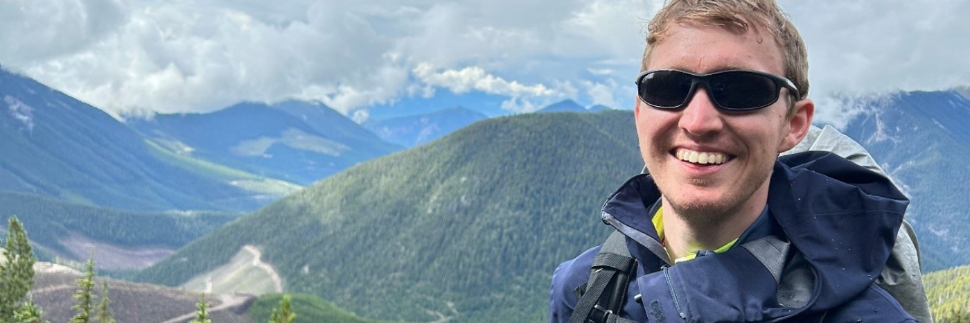 A young man is standing outside on a hill with mountains in the background. He is wearing black sunglasses, a blue jacket with a backpack and has a cane attached to a belt.