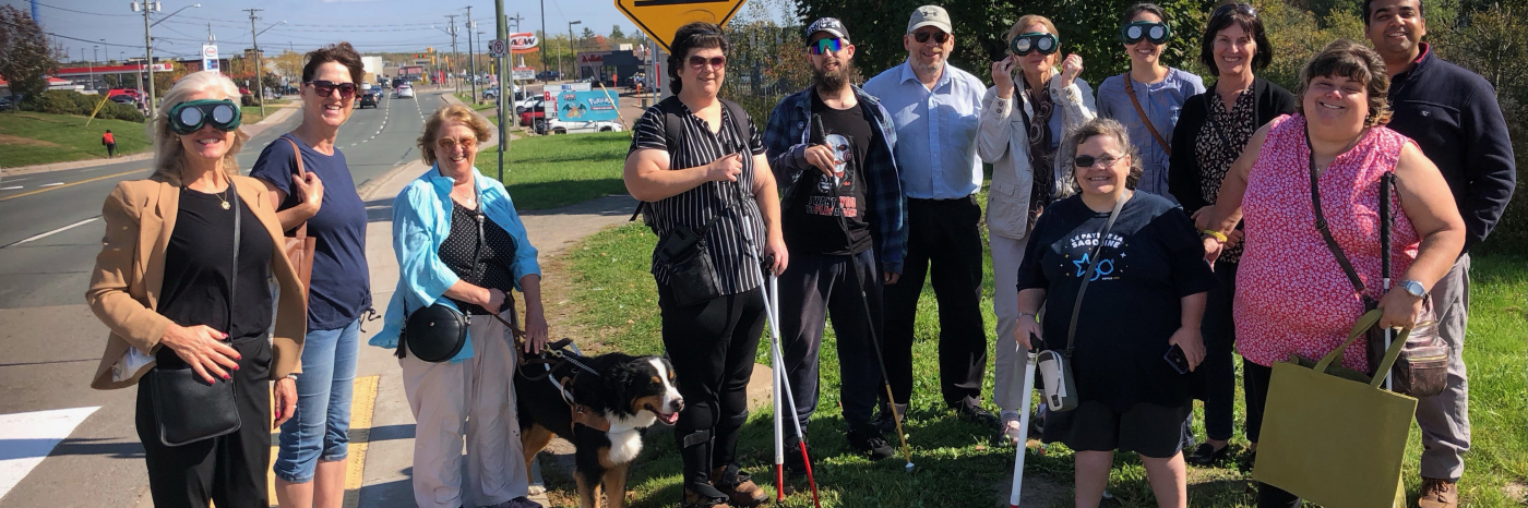 A large group of people stand beside a crosswalk nest to a large road, all smiling. One of the people has a guide dog, several others are wearing glasses that simulate different experiences of blindness. 