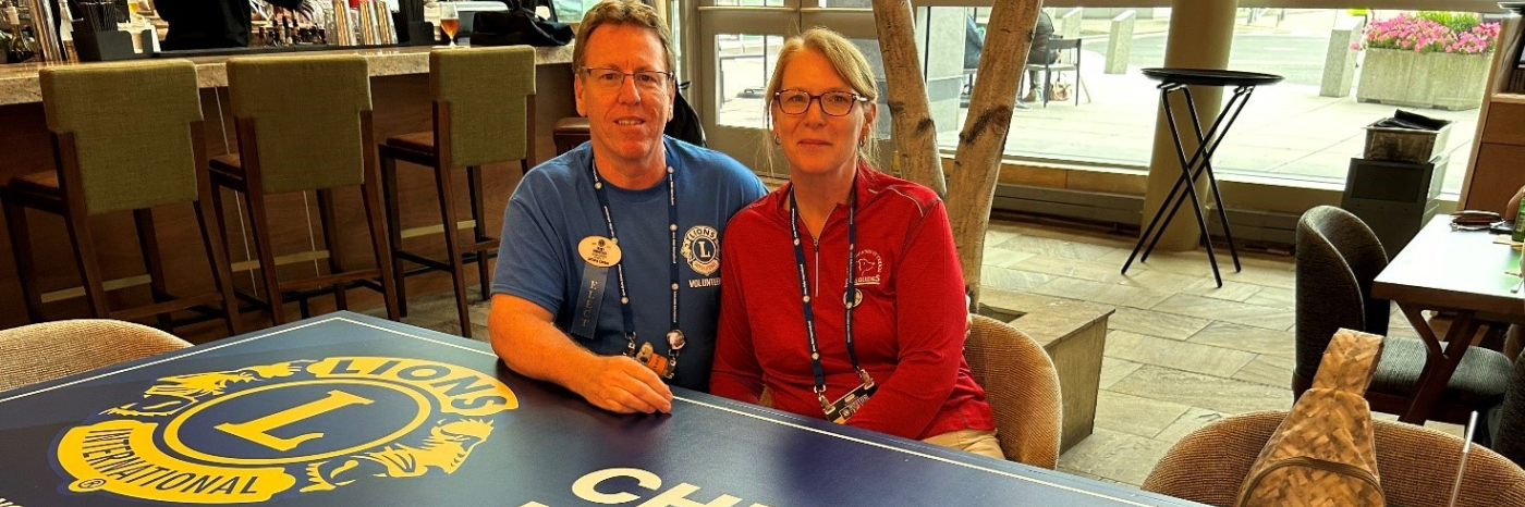 Jerry and Jenn are sitting together behind a table with the Lions International logo and the words “Cheers to Kindness” printed on it.