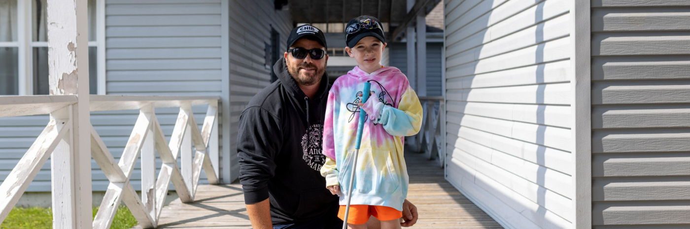 A child stands on a wooden porch walkway between two buildings at CNIB Lake Joe on a sunny day. The child is holding a white cane and is wearing a tie-dye hoodie, bright shorts, sneakers, and a baseball cap with sunglasses resting on it. An adult is kneeling beside the child, wearing a dark hoodie, shorts, sunglasses, and a cap.