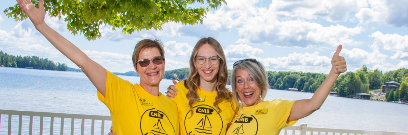 Monique Pilkington, Emma Van Dyk, and Sherri Helsdingen stand together by the Lake Joe waterfront, wearing yellow Lake Joe T-shirts with their arms around one another. They smile at the camera and give thumbs-up gestures.