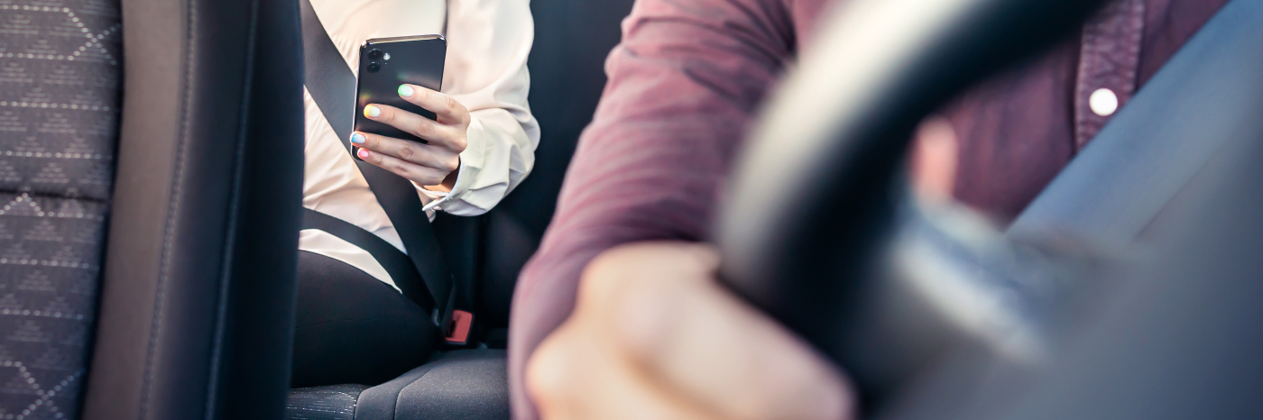 A passenger wearing a seatbelt sits in the back seat of a rideshare car, holding a smartphone while the driver steers from the front seat.