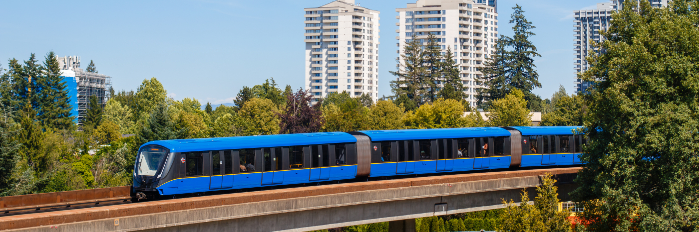 A Vancouver SkyTrain car crosses an elevated track above a tree-filled neighbourhood with residential high-rise buildings under a clear blue sky.