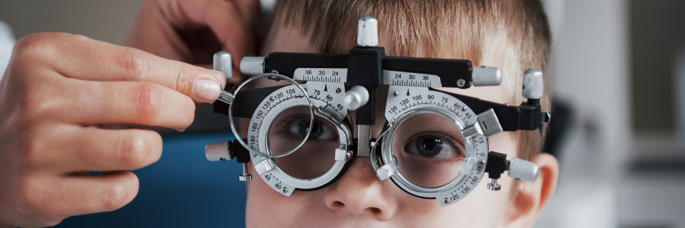 A young child wears a phoropter during an eye exam while an eye care professional adjusts one of the lenses.