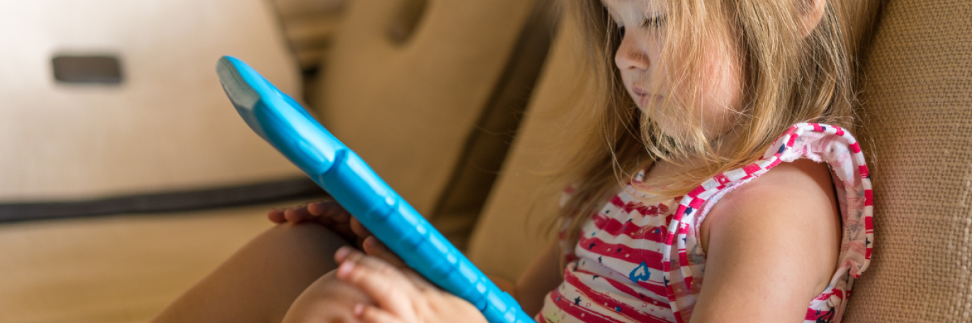A young girl sits on a couch and plays on a tablet. The tablet is in a protective blue rubber case.