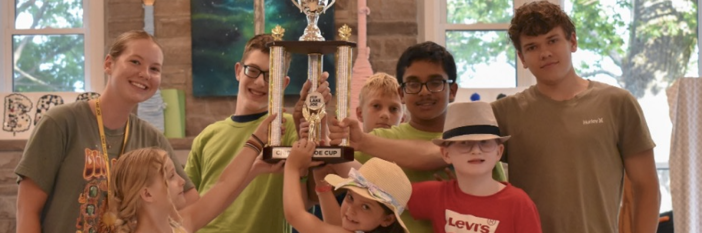 Seven children and youth gather around a large trophy in lounge at CNIB Lake Joe, smiling as they hold it up together. The trophy features a silver cup above three tall gold columns.