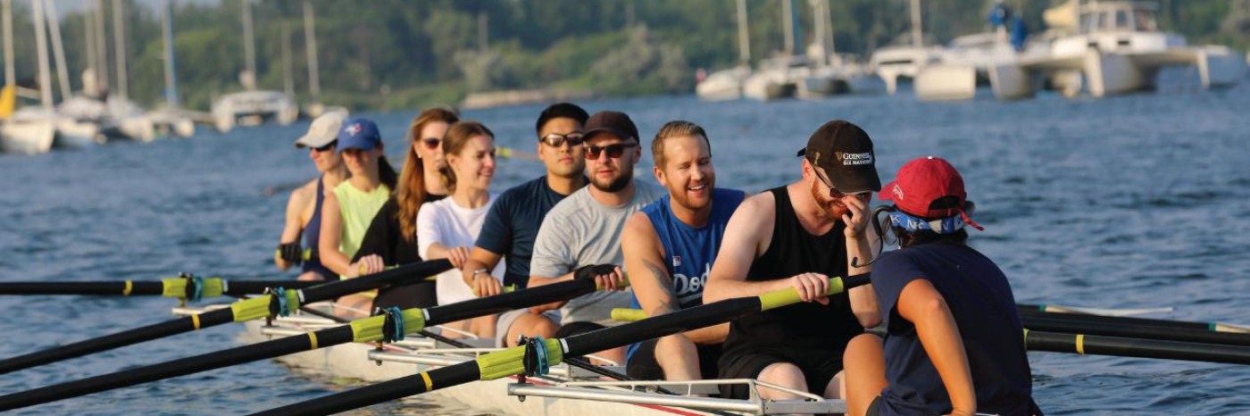 A group of eight people sit in a long rowing shell on calm water, holding oars in position. They appear relaxed and smiling, as if pausing between strokes. In the background, several sailboats are anchored near a tree-lined shore.