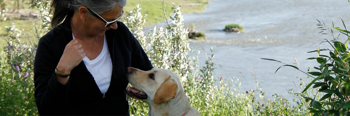 Debra Williamson agenouillée à côté de Patsy, un Labrador-Retriever blond portant un gilet de futur chien-guide, qui s’échangent un regard devant la Bow River à Calgary