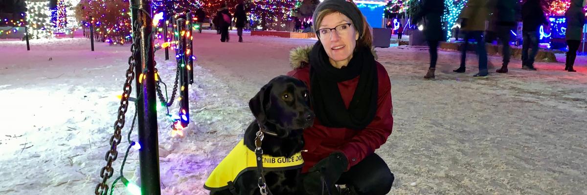 Lorraine Rempel agenouillée sur le sol enneigé à côté de Jennie, un croisement de Labrador-Retriever noir portant un gilet jaune de futur chien-guide. Des arbres ornés de lumières campent le décor derrière eux.