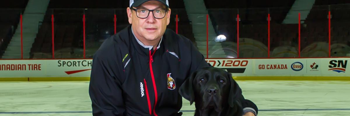 Mark Kelly kneeling next to Rookie, a Future CNIB Guide Dog wearing an Ottawa Senators jersey, with his arm around Rookie while they both smile for the camera. They are on a red carpet laid over the ice of the Canadian Tire Centre, home to the Ottawa Senators.