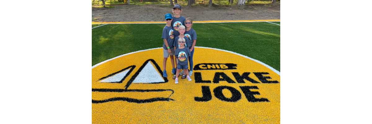 Five children posing on the giant yellow CNIB Lake Joe logo in the middle of the new soccer field. They are all wearing grey CNIB Lake Joe tshirts.