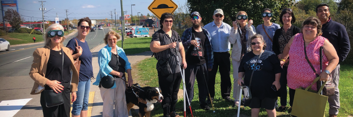 Un groupe de personnes ayant une limitation visuelle se tient à côté d'un passage pour piétons avec des agents publics, à Moncton lors de la campagne « Montez à bord » de l'automne 2023.