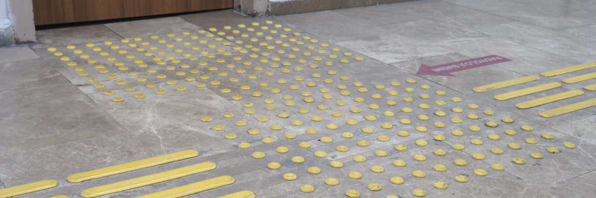 A tiled floor with tactile paving/ tactile wayfinding surface indicators. The paving consists of yellow raised dots and lines. 