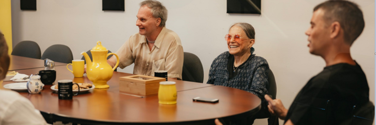 Four people sit around a boardroom table, laughing and talking, with a teapot, mugs, and snacks.