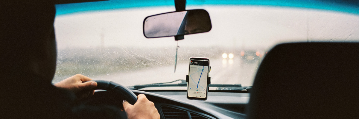 Inside a rideshare car, the photograph is taken from the passenger’s point of view in the back seat. There is a smartphone mounted on the dashboard of the car displaying a rideshare app. The driver’s hand is on the steering wheel and it is raining outside. 