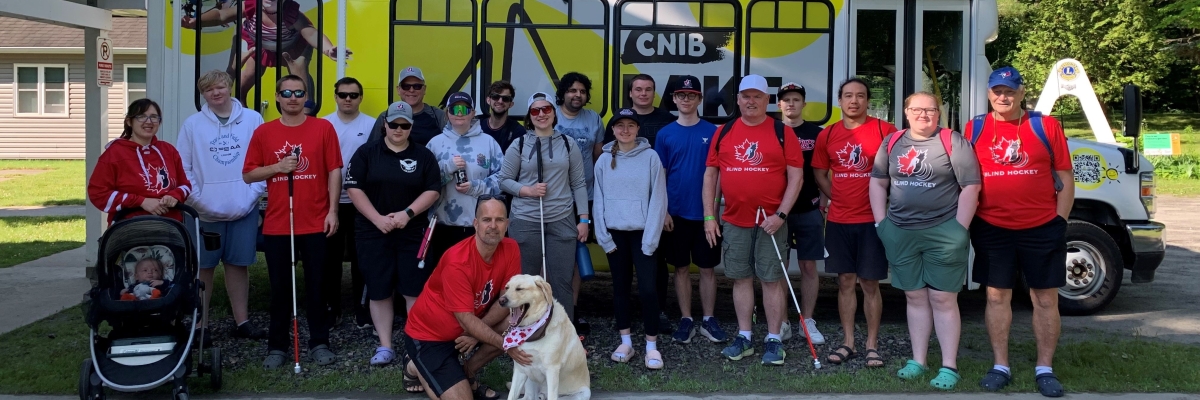 Members of the Canadian Blind Hockey Association (details) pose in front of the minibus at the CNIB Lake Joe Welcome Centre. They are wearing XX hockey jerseys and holding big hockey bags.  