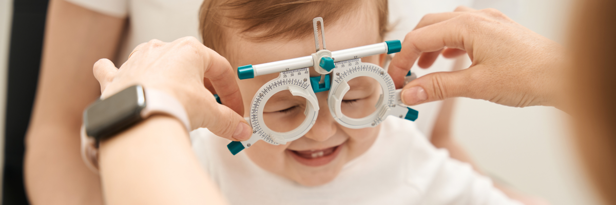 A baby is smiling while having their eyes examined. An adult is holding a measurement device up to the baby’s face.