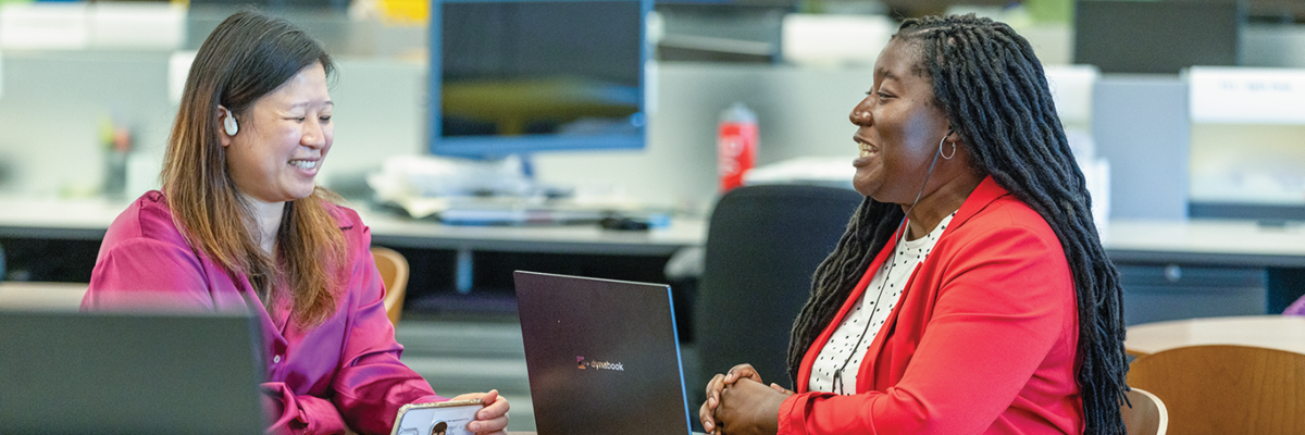 Two women sit at a table in an office environment, engaged in a conversation. Both have laptops in front of them on the table. In the background are computers and different workstations.
