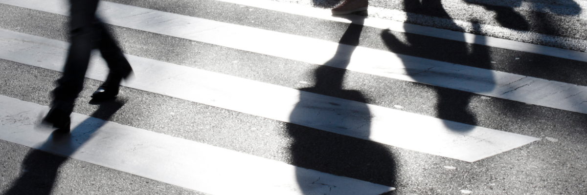 A crosswalk with shadows of people walking. The image is in black and white, showing the lower valves of several people as they cross a street. 