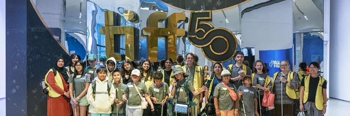 In the lobby of the TIFF Lightbox, a large group of young campers and CNIB volunteers pose for a group photo in front of a giant “TIFF 50” sign.