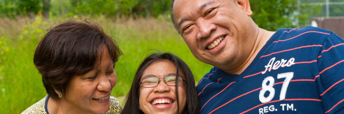 A mother, father, and teenage daughter laugh together; the daughter is wearing glasses.