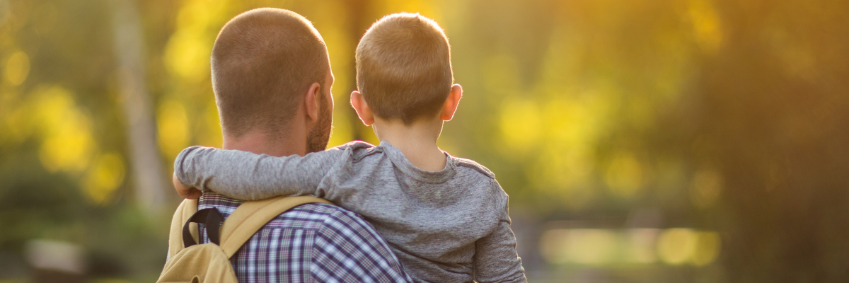 A father holds his young son. The photo is taken from behind, and the child has their arm around their father's shoulders. The background is a blurry outdoor scene with yellow and green tones.