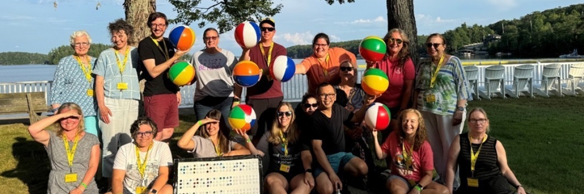 A group of CNIB Lake Joe staff and volunteers smiling outdoors on a sunny day by Lake Joseph. They are holding colourful beach balls printed with “BRL” to represent the word braille. They are posing under tall trees with white chairs and water in the background.