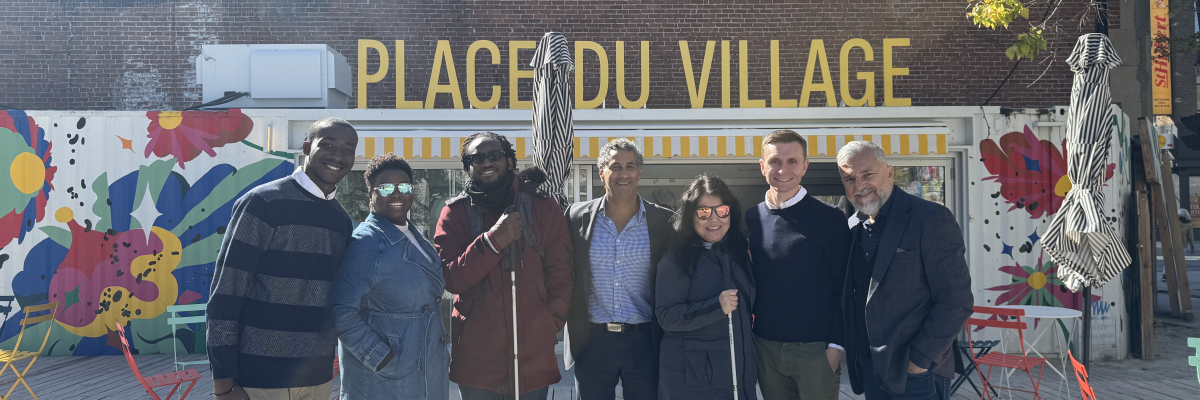 Shalom Foly, Thama Cherilus, Eitel Houédakor, Jean-Pierre Khouzam, Najla Noori, Chris McCray et Cosimo La Rosa posent devant la Place du Village, sous le soleil. Eitel et Najla tiennent leur canne blanche.