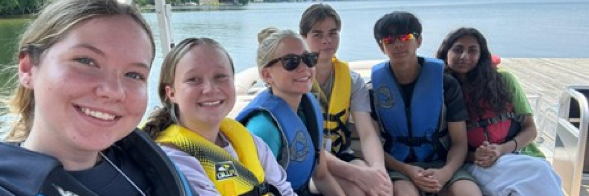 Young men and women are on a pontoon boat on the lake wearing colourful life jackets.
