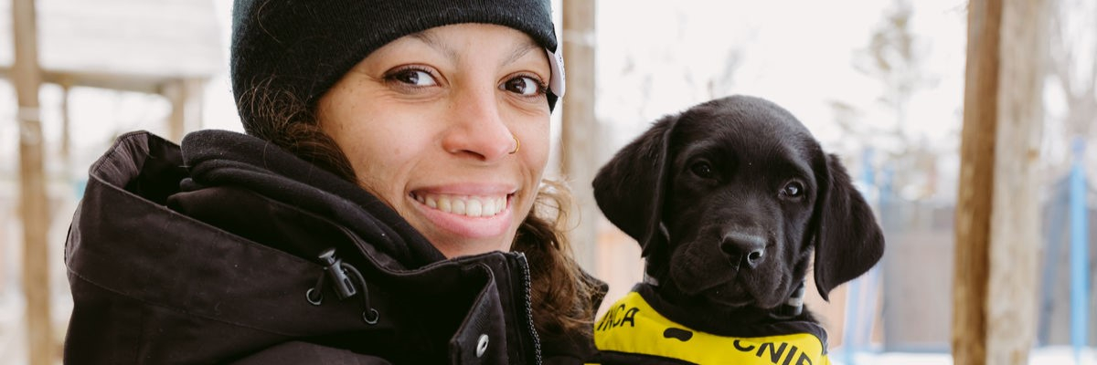 A smiling person in a winter jacket holds a black puppy wearing a CNIB Future Guide Dog vest.