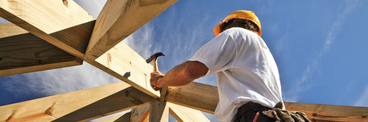 Construction worker wearing a hard hat uses a hammer to secure wooden roof beams, viewed from below against a bright blue sky.