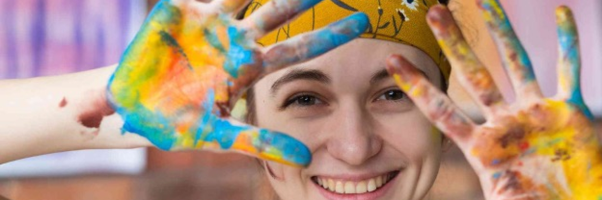 A young woman is smiling at the camera. Her hair is pulled back by a gold flowered bandana. She is holding her hands open in front of her, and they are covered in bright paint colours.