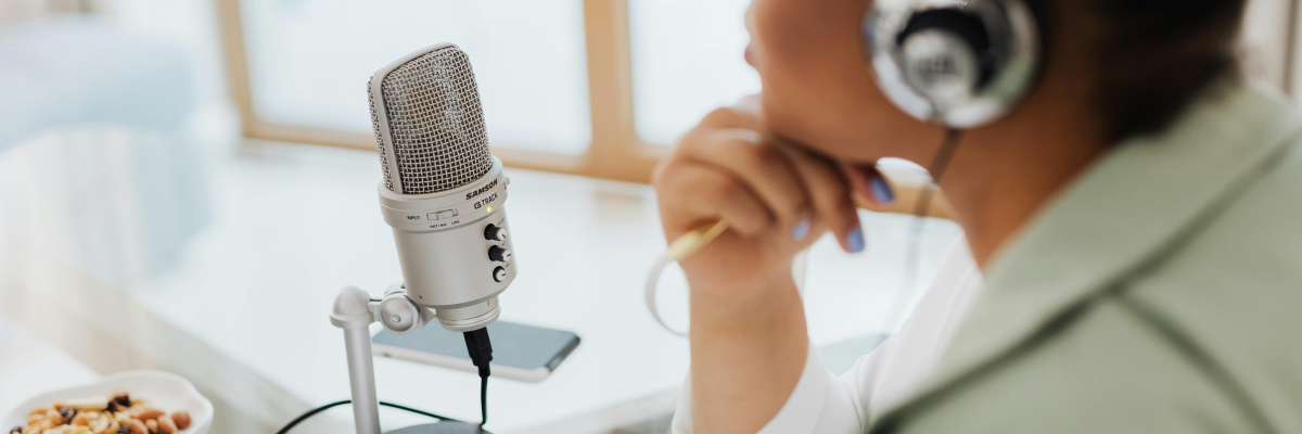 A woman wearing over-ear headphones sits at a desk, positioned beside a microphone on a stand. They are recording a podcast. 