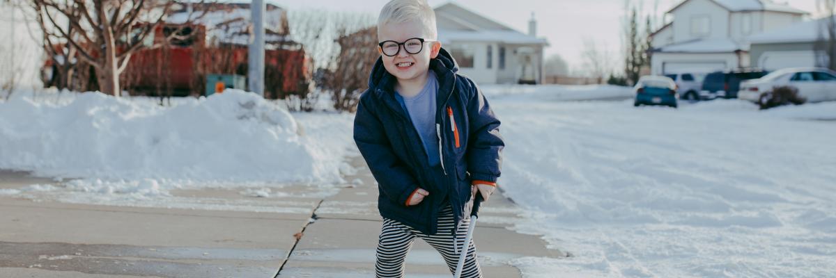 Little boy on a sidewalk with snow around him, holding his white cane like a hockey stick. He's smiling.