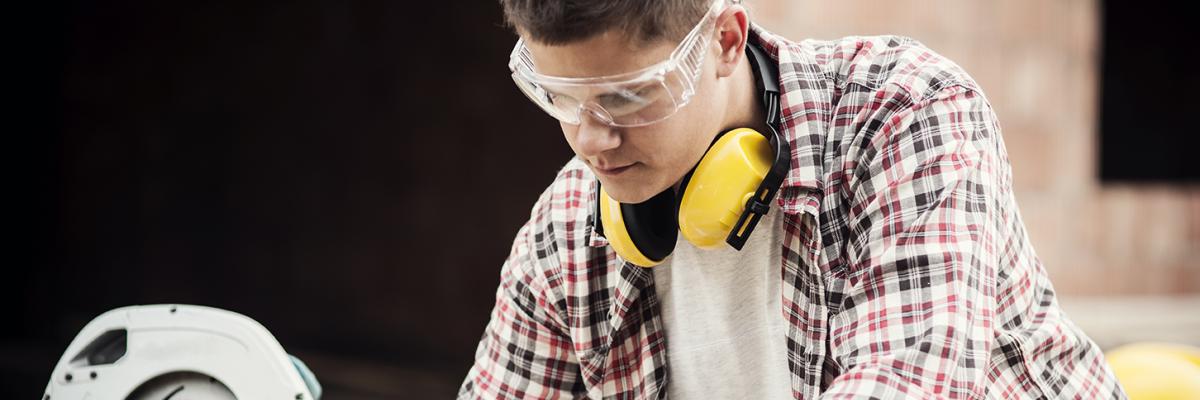 Man looking down and marking a piece of wood with a pencil. He is wearing protective glasses.