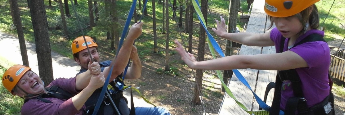 A man and a woman are leaning back attached to harnesses with their feet braced against a ziplining platform. Their daughter stands on the platform harnessed with her arms reaching toward them