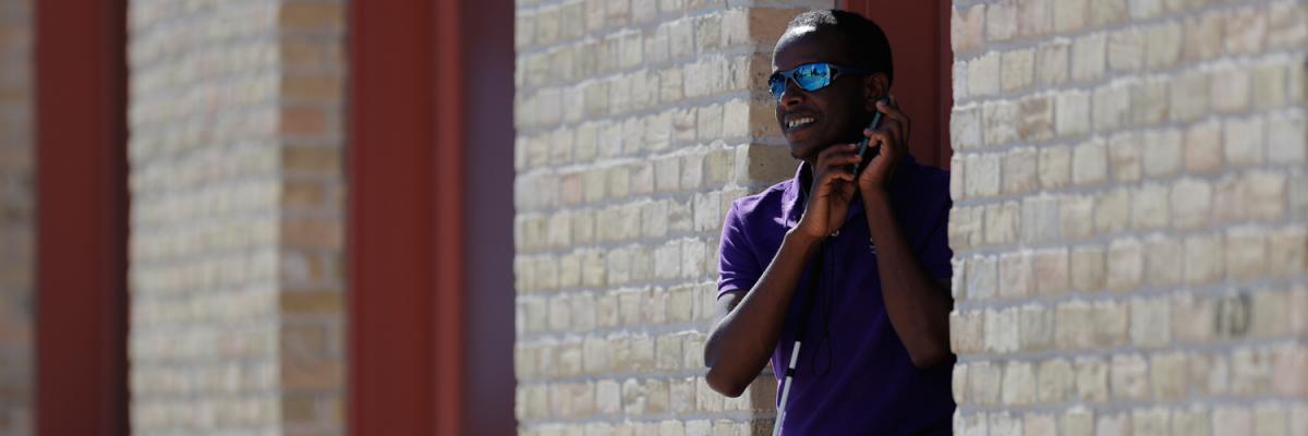 A man holds an iPhone up to his ear.  He is wearing a purple shirt, sunglasses and is standing outside against a brick wall. 
