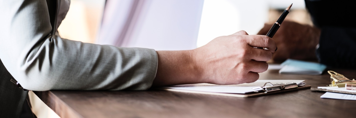 Close up of a person writing on a piece of paper with another person across the table from them