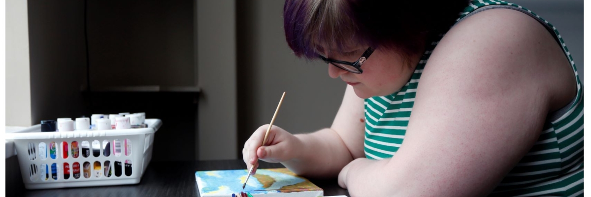 A young girl wearing glasses, painting on a canvas resting on a table.