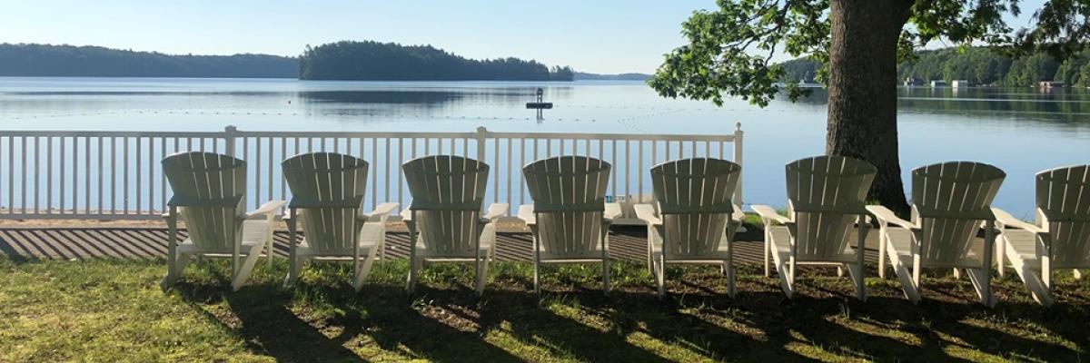 A row of white, Muskoka chairs sit on the shore of Lake Joe. 