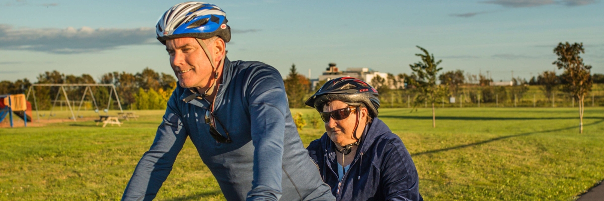 A man and woman riding a red tandem bike through the park on a sunny day.