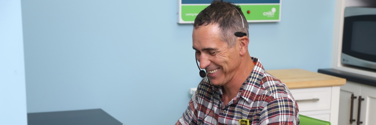 A smiling man wearing a telephone headset takes notes on a clipboard.