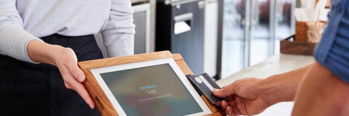 Description de la photo : Une personne hors cadre passe sa carte bancaire sur l’écran tactile d’un terminal de paiement qui lui est présenté par l’employée d’un café.