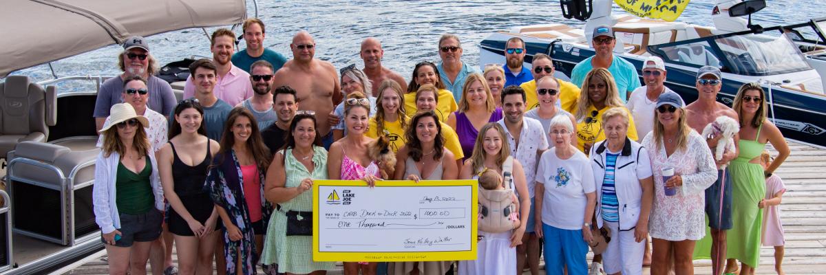 A group shot of the partygoers at the Dock to Dock event. Together they hold a large cheque. 