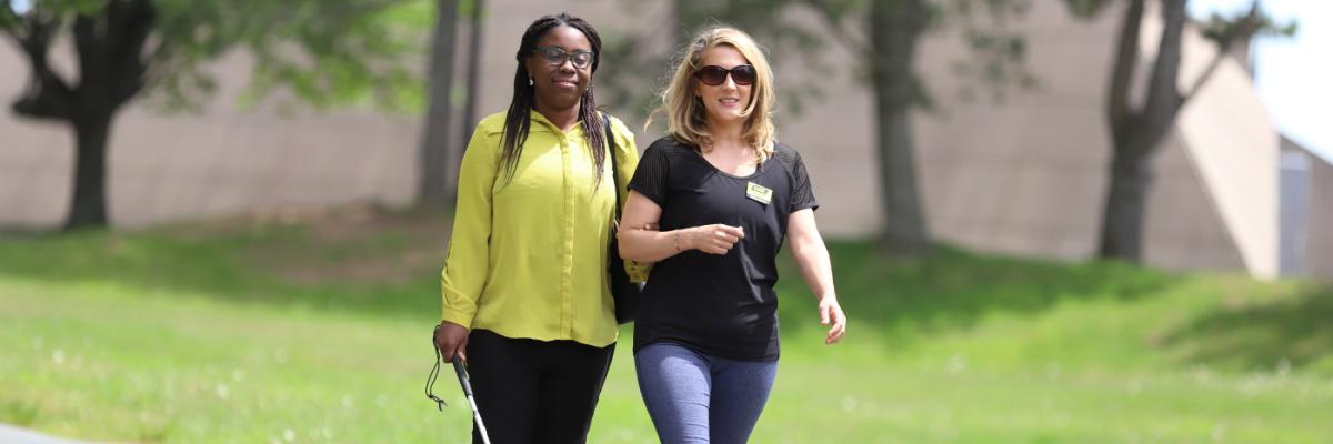 A woman provides sighted guide to another woman who is navigating with a white cane