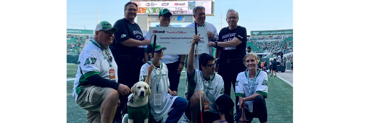 Representatives from CNIB and the Brandt Group of Companies pose together for a photo while holding the $2M cheque at the Saskatchewan Roughriders Game. There are two CNIB Guide Dogs wearing green jerseys. 