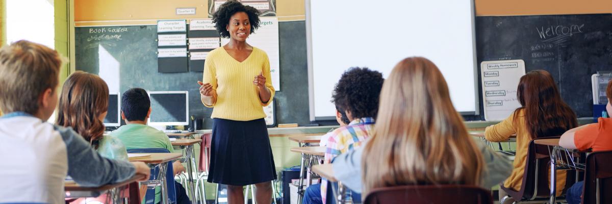 A young woman stands at the front of a classroom and speaks to a group of young students. 